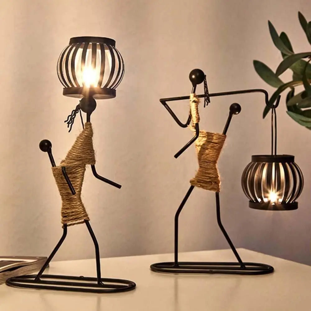 Two abstract figure metal candle holders, one holding a lantern and one with a lantern on the head, softly lit on a tabletop