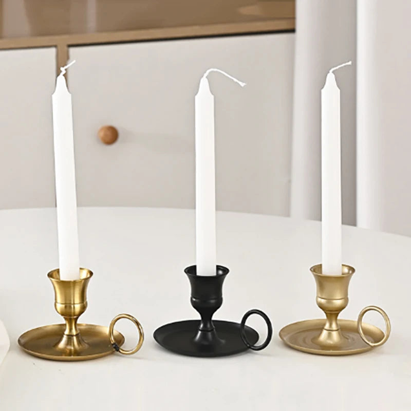 Three chamberstick-style candle holders—gold, black, and light gold—each holding a white taper candle, arranged on a white table