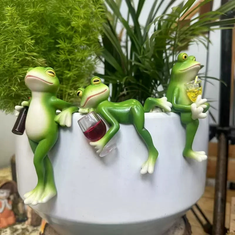 Three resin frog figures perched on the edge of a white planter with green plants, each holding a drink and showing different playful poses