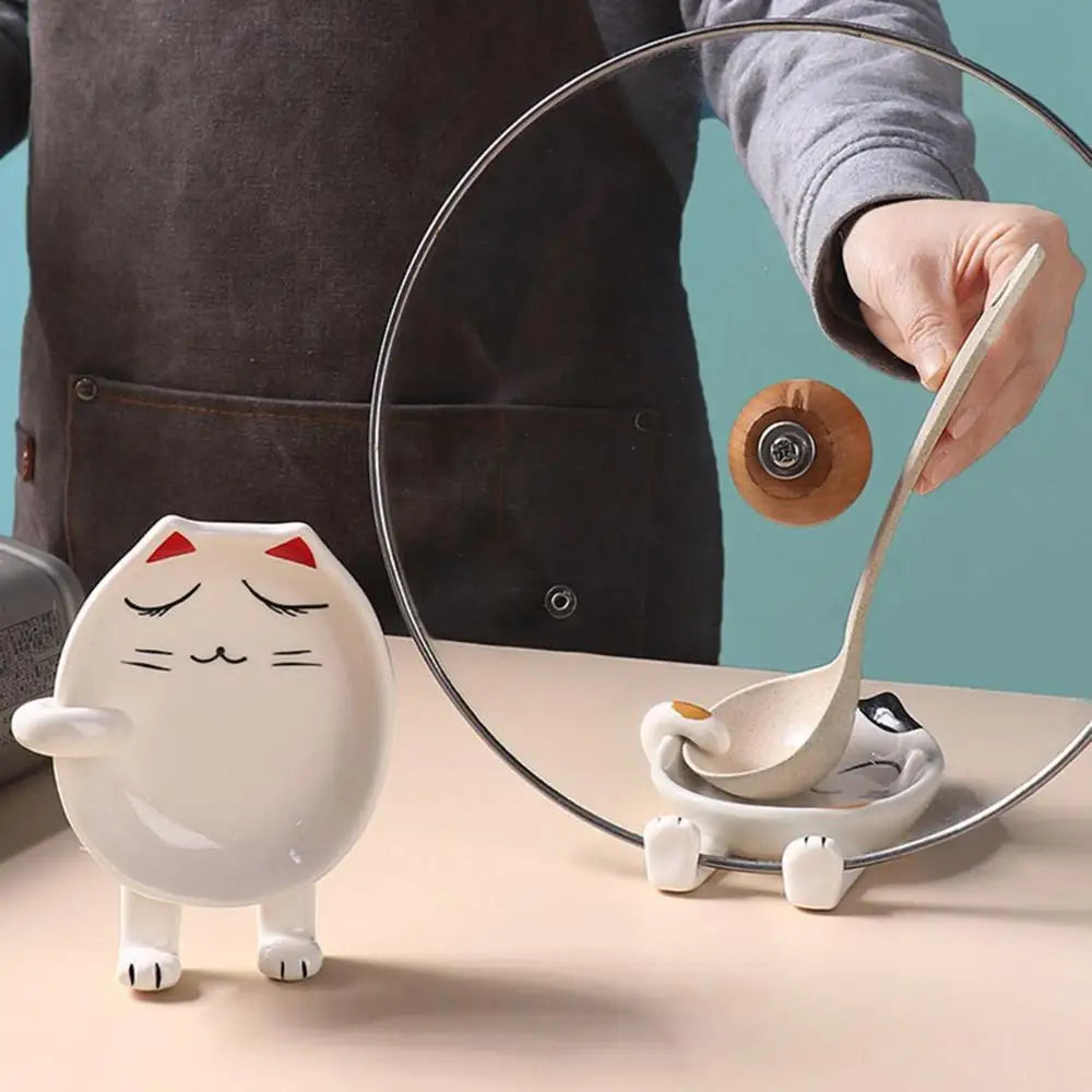 Standing cat-shaped ceramic spoon rest next to a spoon rest in use with a pot lid and ladle on a counter