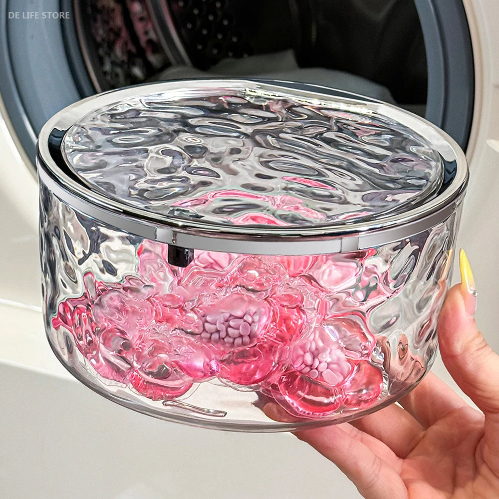 Clear acrylic storage container with pop-up lid holding pink laundry pods near a washing machine