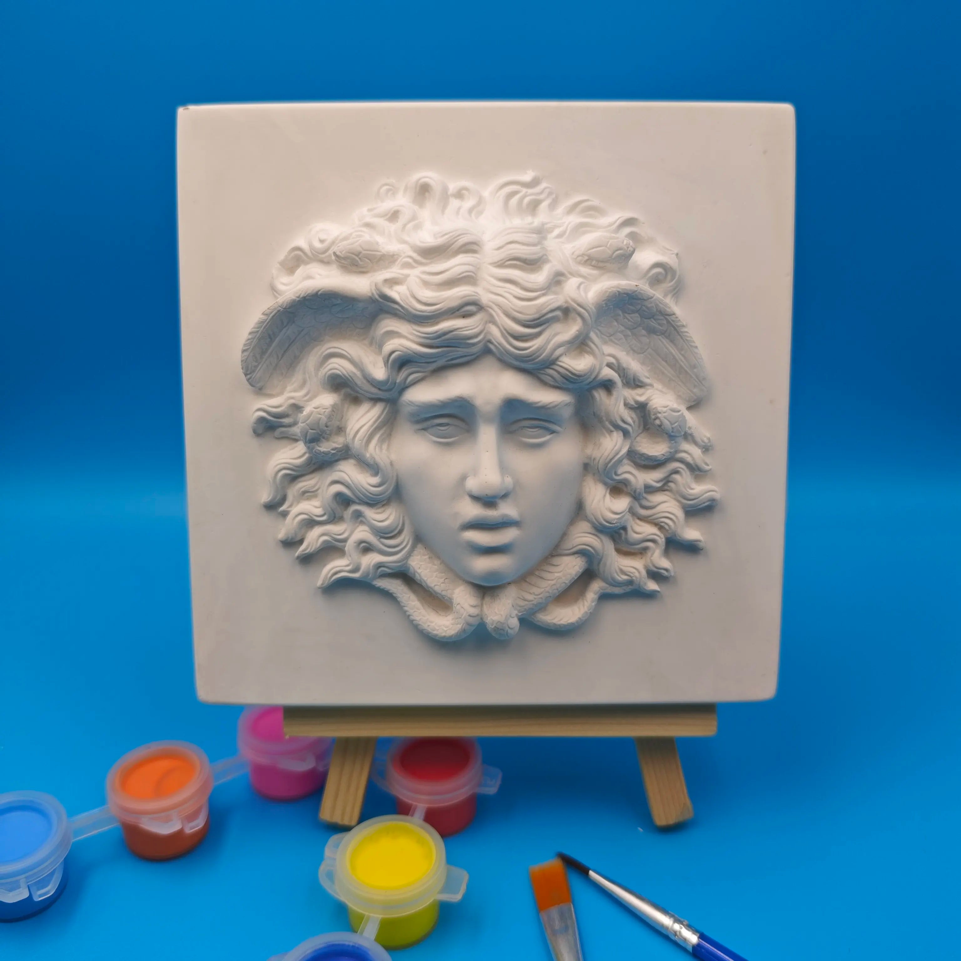 white square Medusa relief wall sculpture with paint pots and brushes on blue background
