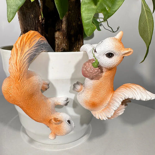 Two orange and white resin squirrel figurines hanging playfully from the edge of a white plant pot