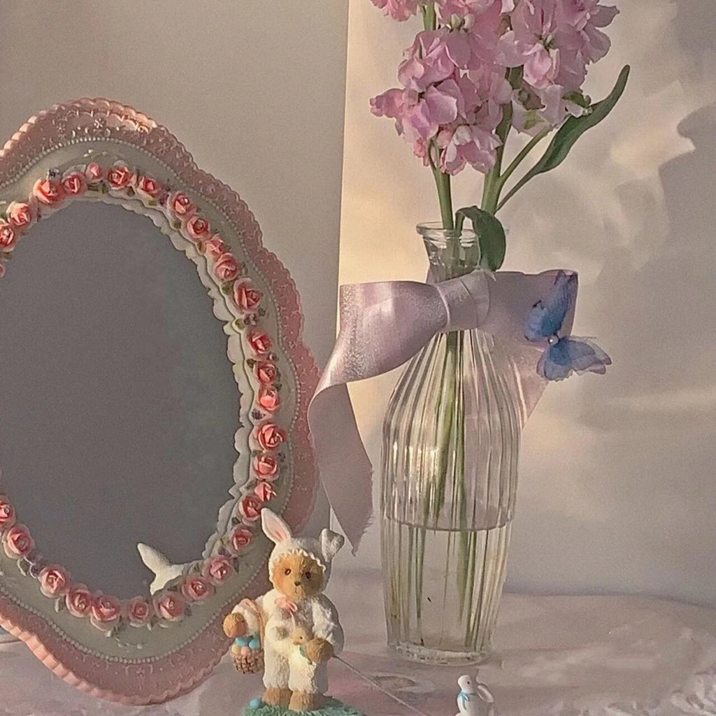 Pink oval desktop mirror with rose border on a vanity table, next to a vase of flowers