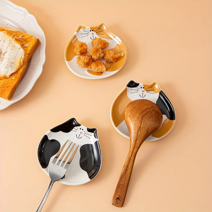 Three cat-shaped ceramic spoon rests in different colors with utensil and snack display on a peach countertop