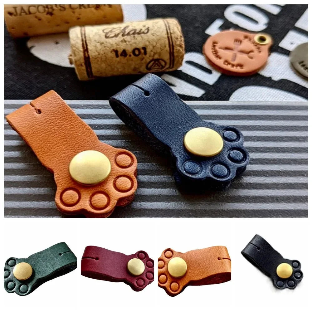 Lineup of cat paw leather cable winder clips in dark blue, brown, green, and wine red colors on a table